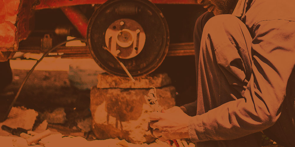 Man in mechanic's uniforn, kneeling next to tireless car on a cement block, working.